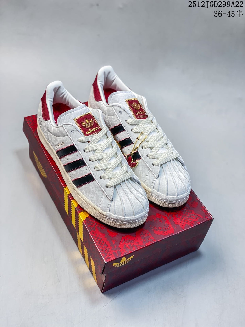 Pair of white Adidas Superstar sneakers with black and red side stripes, red details, and a red patterned box