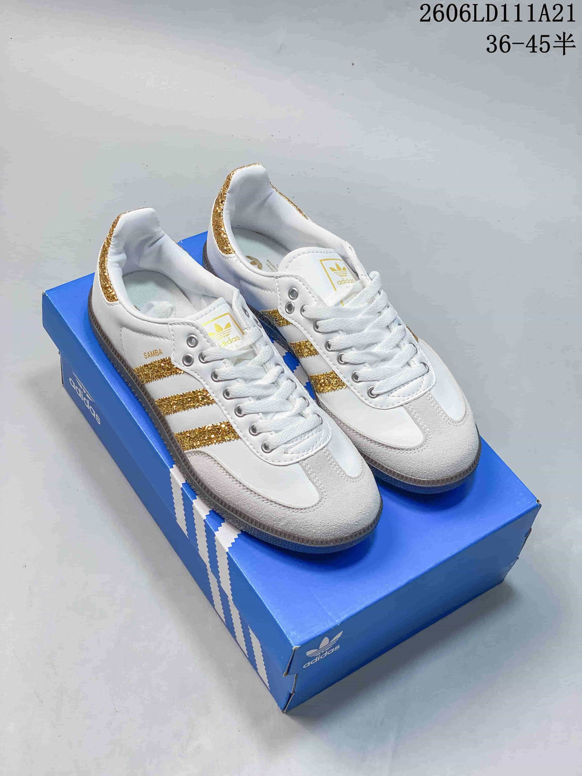 White low-top Samba-style sneakers with gold glitter stripes and heel accents on a blue shoe box