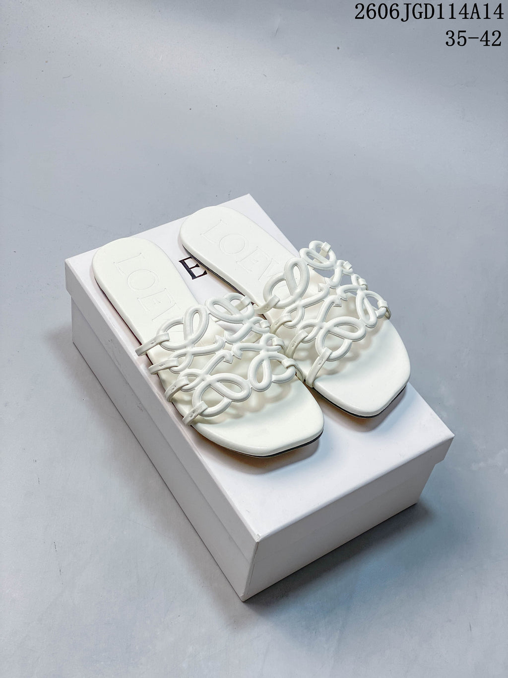 Pair of white Loewe-style flat slide sandals with sculptural interlaced straps, shown on a box