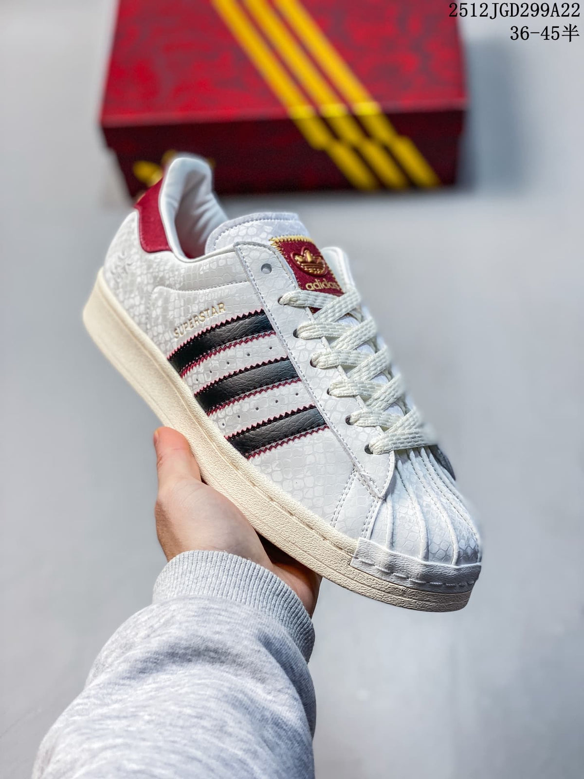 Angled view of a white Adidas Superstar sneaker with textured upper, black and red stripes, and red accents