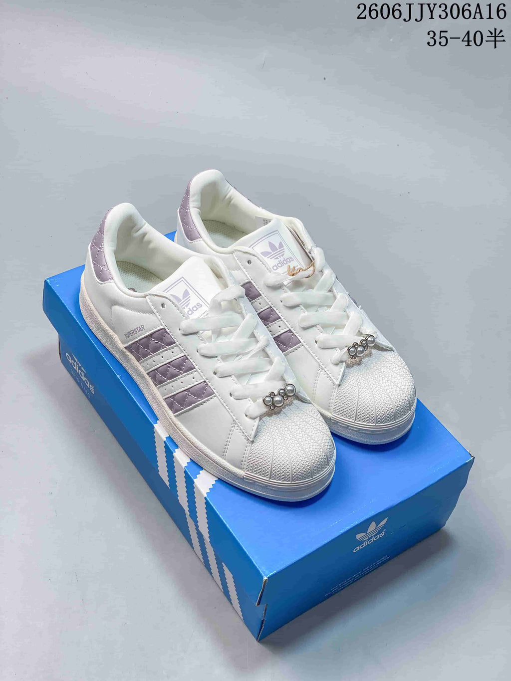 Pair of white Adidas Superstar II sneakers with lilac quilted stripes and heel details, shown on a blue box