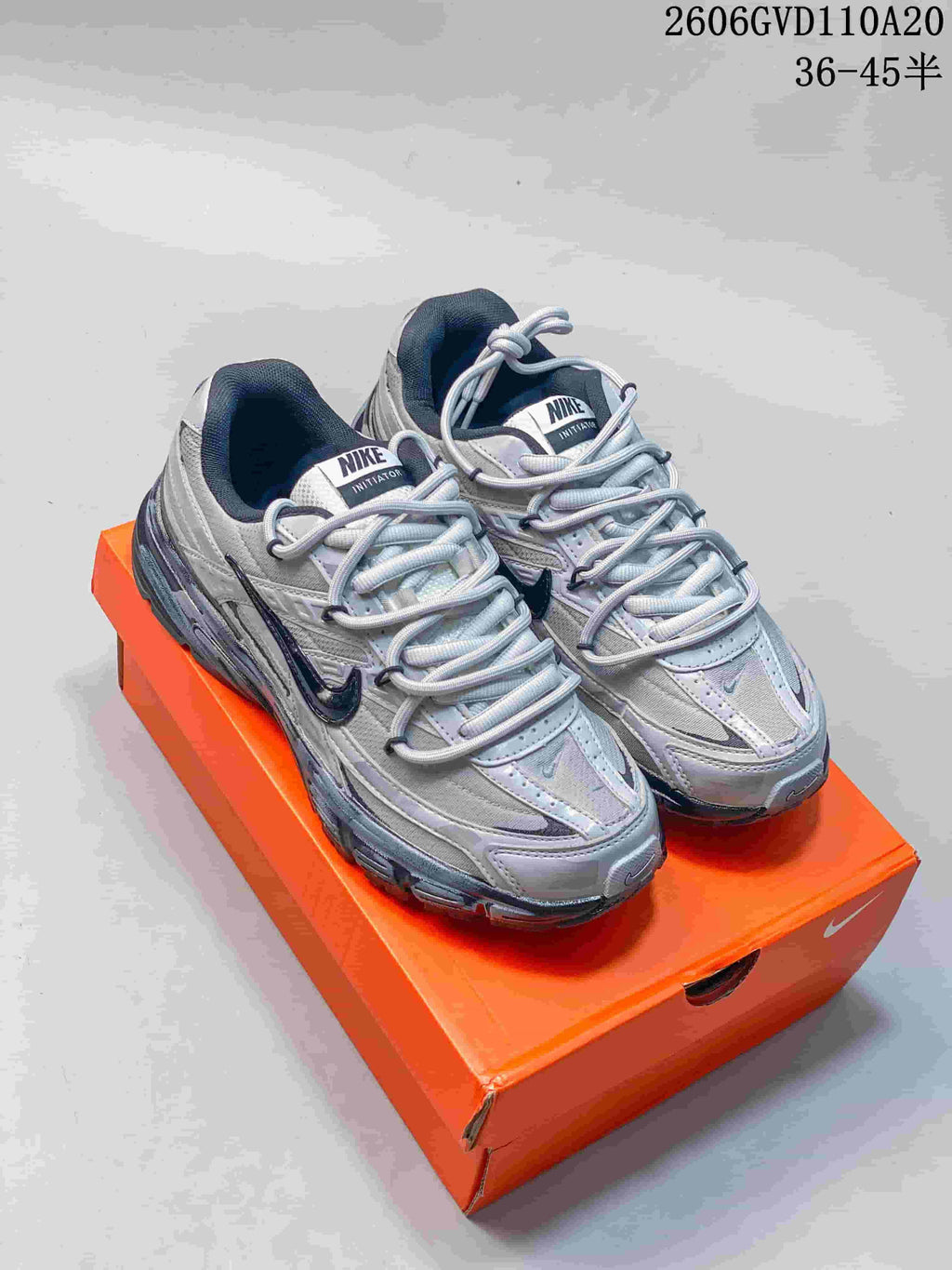Pair of silver and gray Nike Initiator low-top sneakers with dark accents, shown on an orange shoe box.