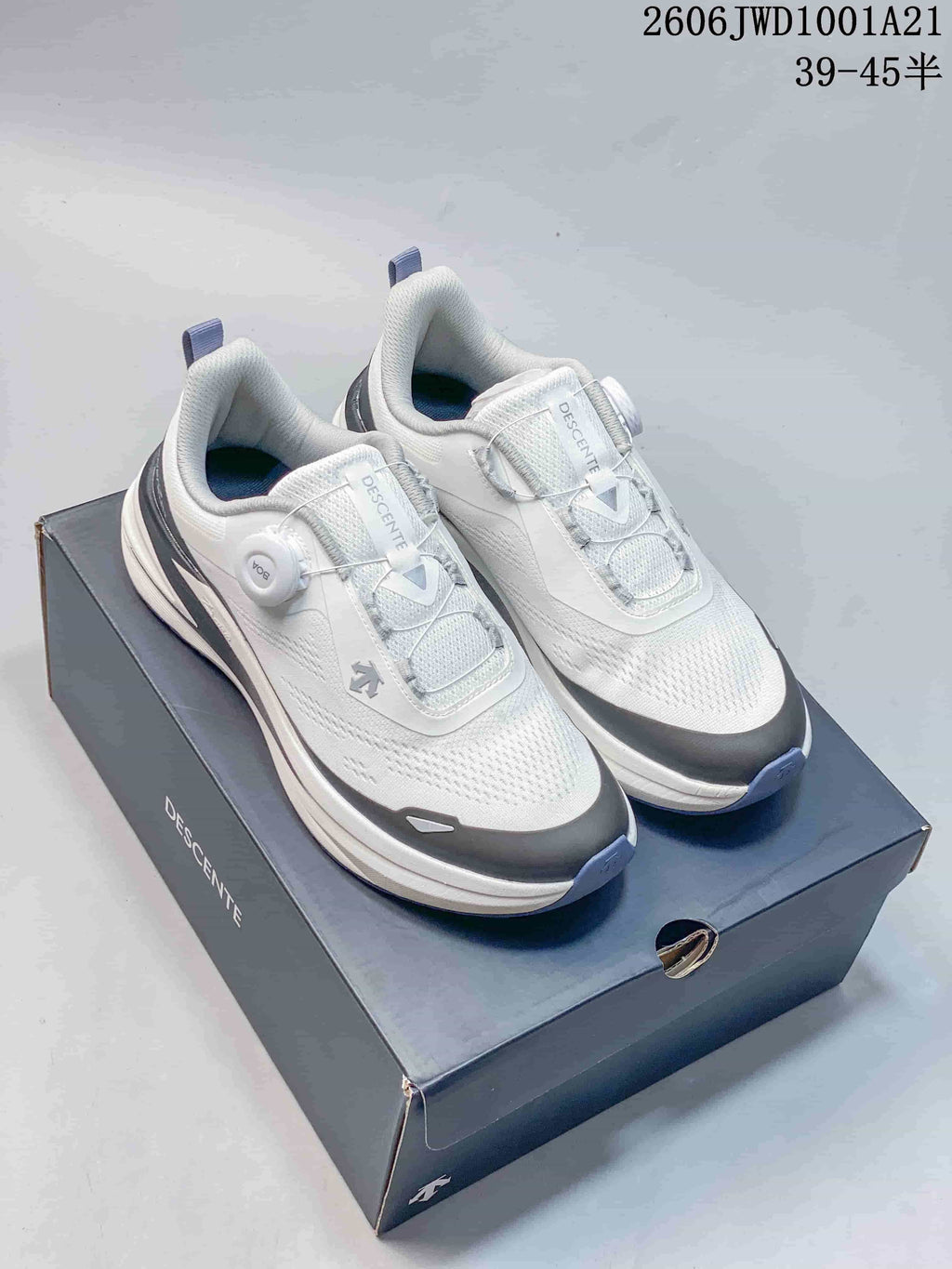 White DESCENTE low-top sneakers with a dial closure, shown on a shoe box; image text lists model code 2606JWD1001A21 and sizes 39–45.