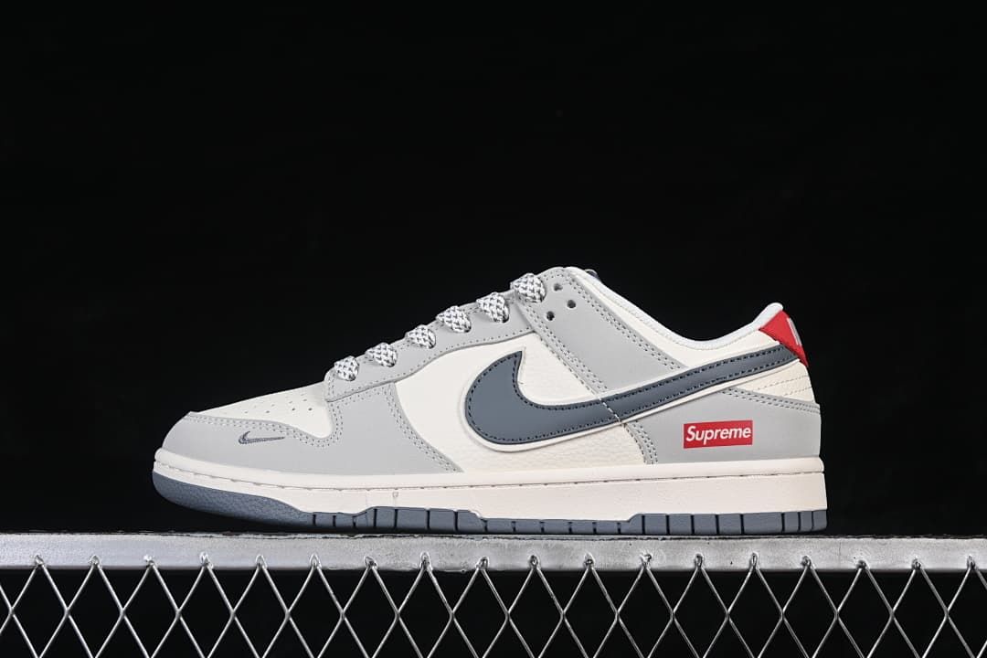 Side view of a grey and off-white Nike SB Dunk Low sneaker with a dark grey Swoosh and red Supreme branding.