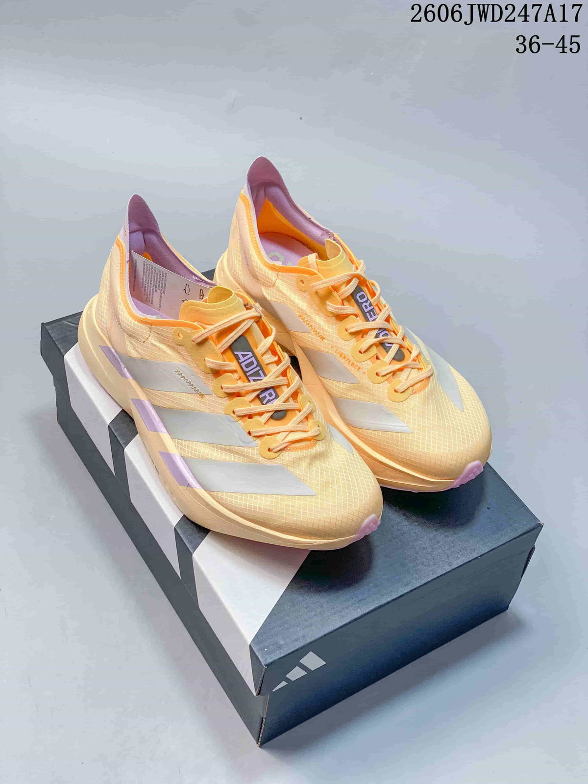 Pair of pale yellow Adidas Adizero running shoes with orange laces and lavender-pink accents, shown on a shoe box.