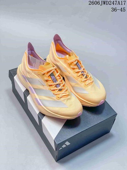 Pair of pale yellow Adidas Adizero running shoes with orange laces and lavender-pink accents, shown on a shoe box.