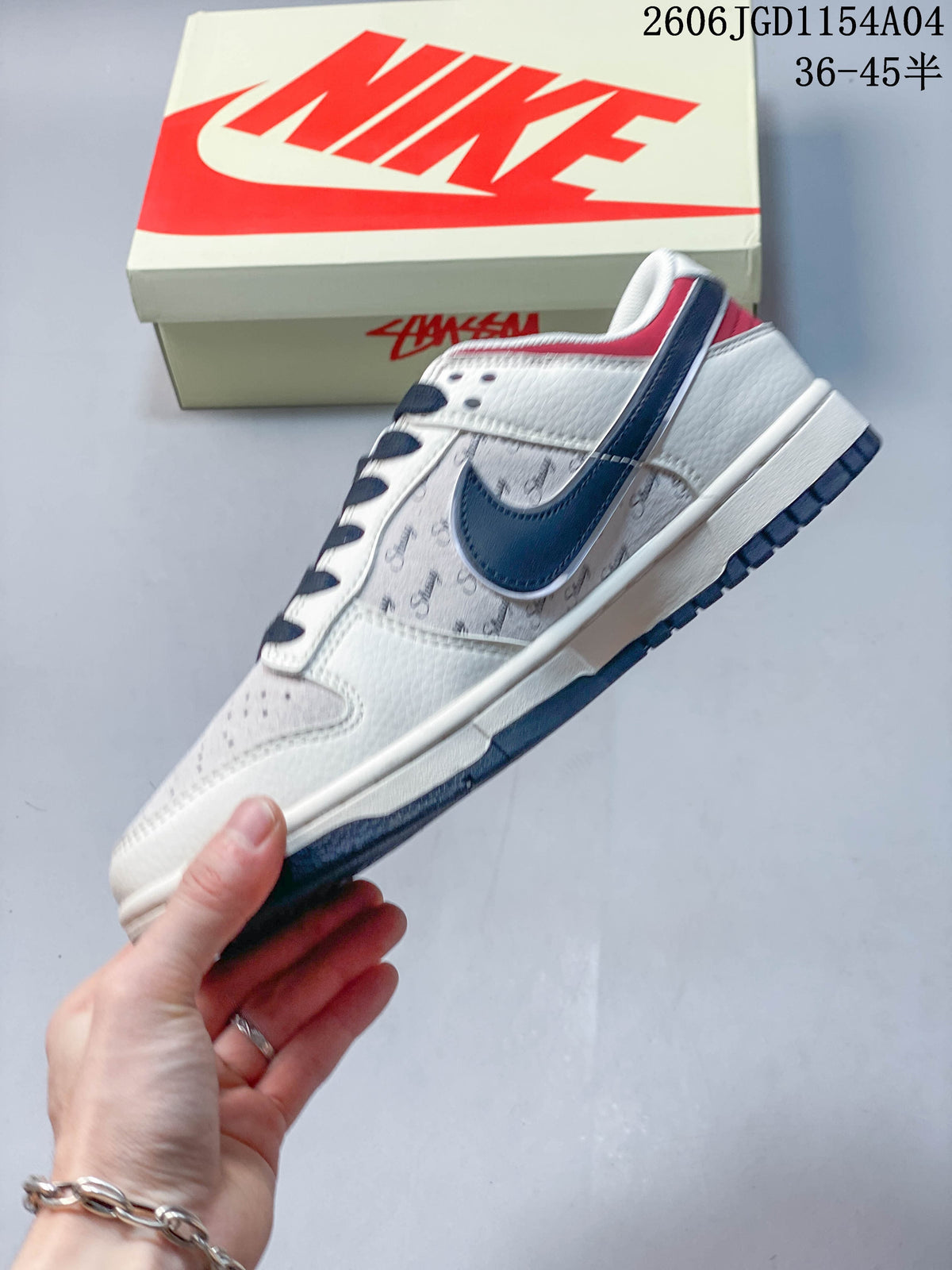 Side view of a light Nike SB Dunk Low sneaker with a navy Swoosh, script-patterned panel, and red heel accent.