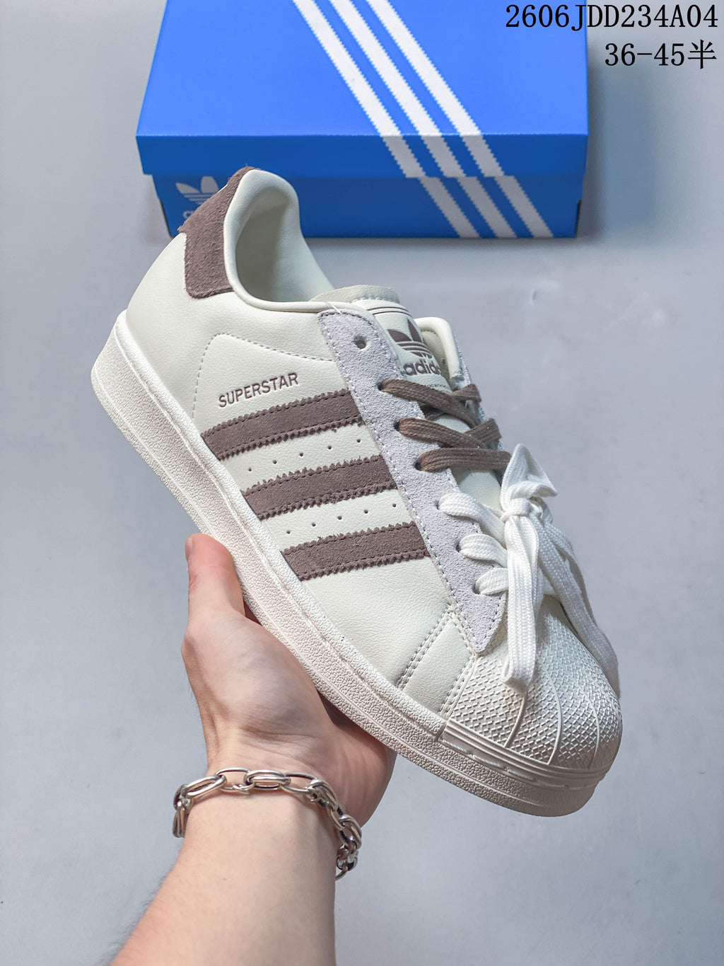 Angled side view of an off-white Adidas Superstar sneaker with brown stripes and a gray overlay.