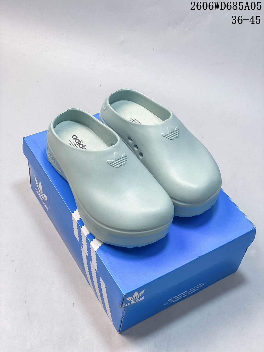 Pair of light blue Adidas AdiFOM Stan Smith mule shoes displayed on a blue shoe box