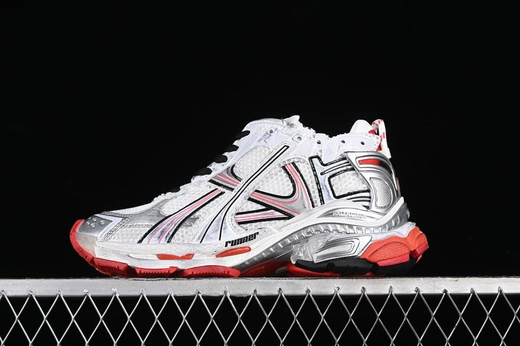 Side view of a white Balenciaga Runner-style sneaker with silver overlays, black detailing and red accents.
