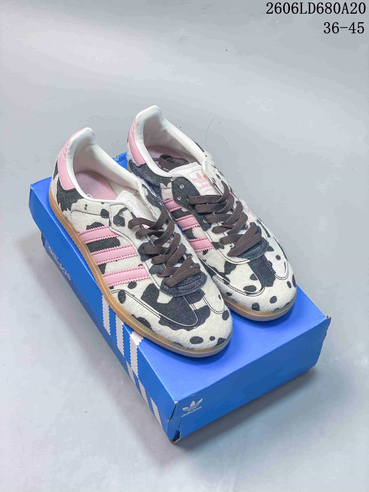 Adidas Samba OG sneakers with cow-print uppers, pink stripes, dark laces, and gum-colored soles on a blue shoebox