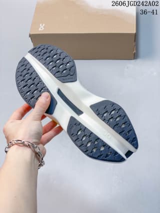 Underside view of the sneakers showing the dark patterned outsole and white midsole, with a shoebox in the background.