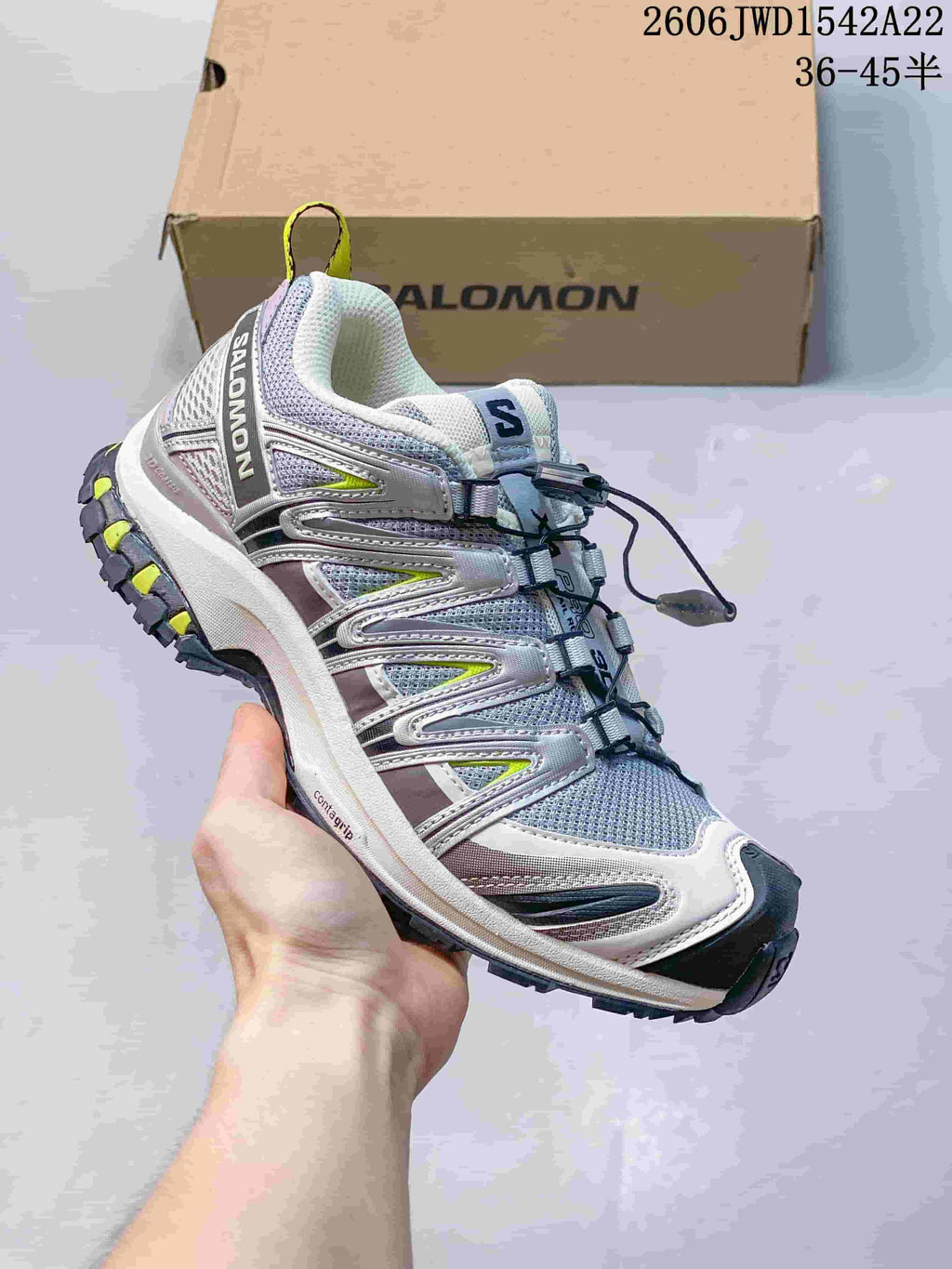 Light gray Salomon trail-style sneaker with reflective-look overlays and bright yellow accents, shown beside a branded box.