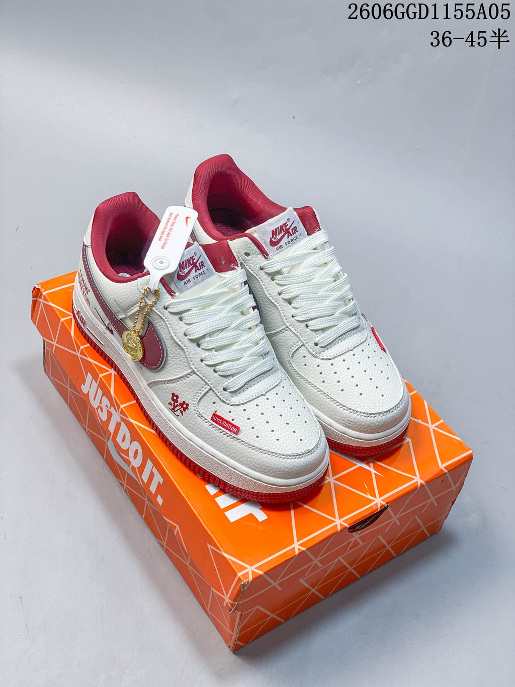 Pair of white low-top sneakers with burgundy-red accents and a gold-tone lace charm, shown on an orange shoe box