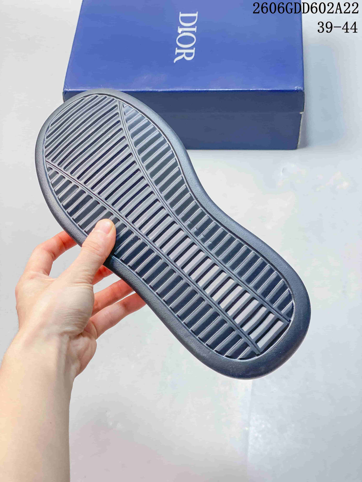 Underside of a sneaker showing a grooved outsole, with a blue Dior box behind it