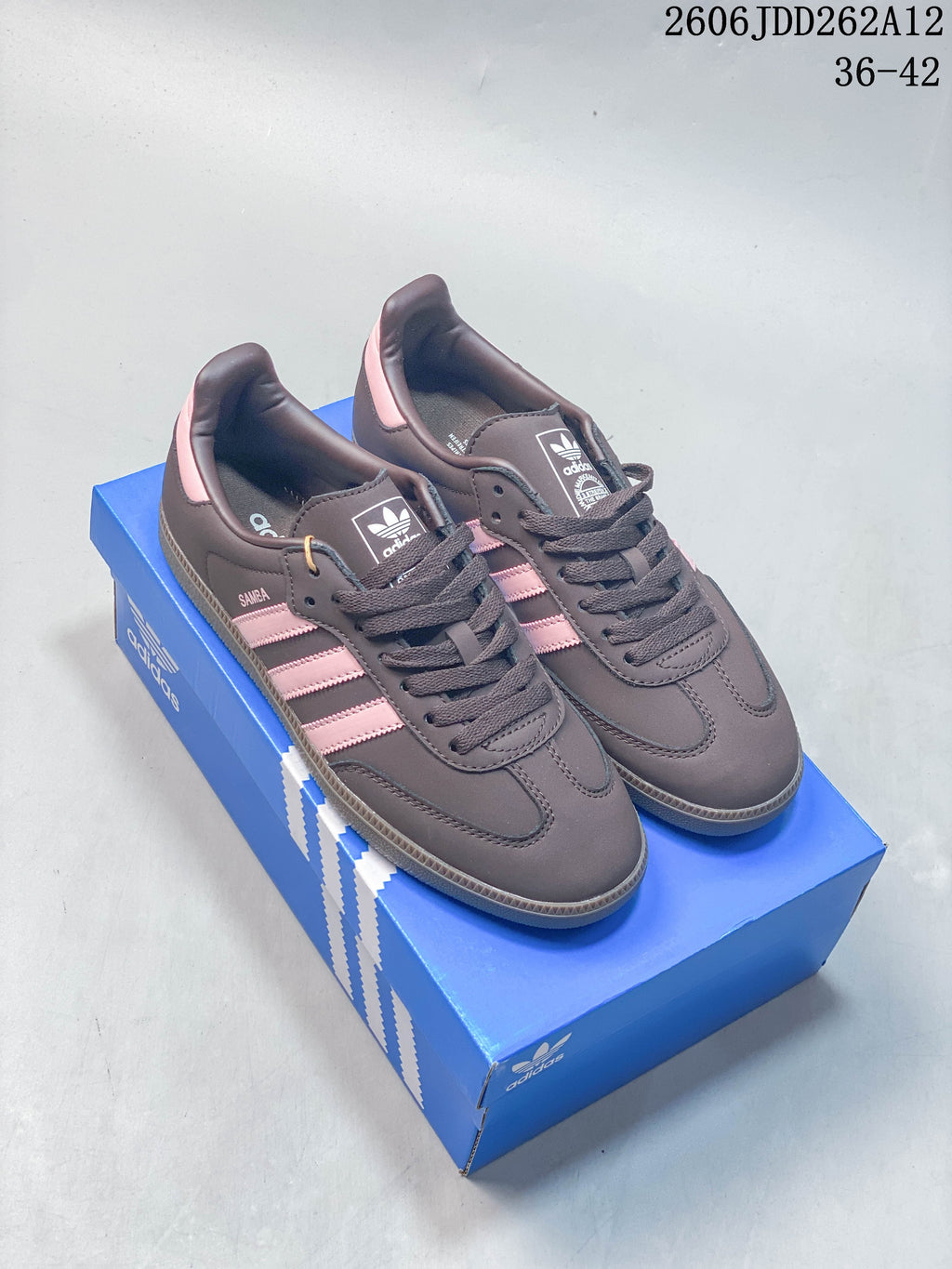 Dark brown low-top Samba-style sneakers with pink stripes beside silver sneakers on a blue box.