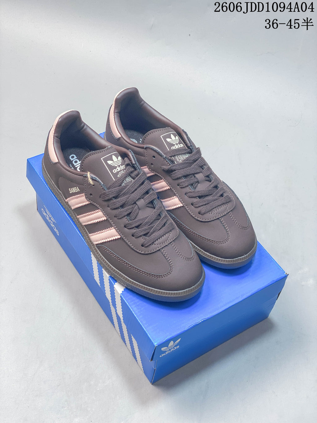Pair of gray-brown Adidas Samba OG sneakers with pale pink stripes displayed on a blue shoe box