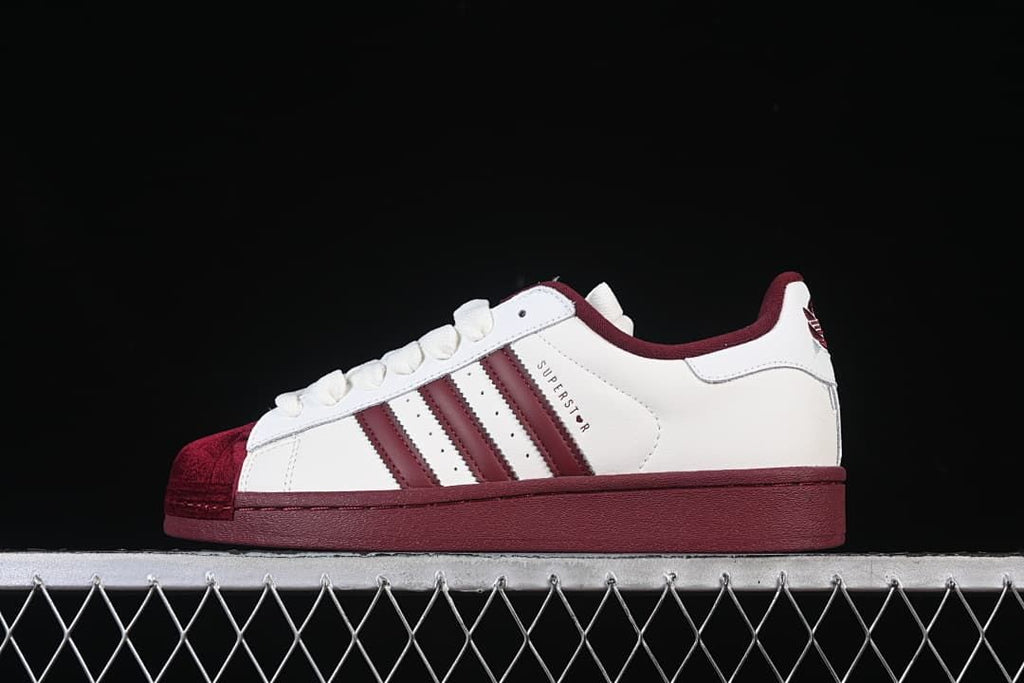Side view of white adidas Superstar II sneakers with burgundy stripes, sole, and shell-style toe.