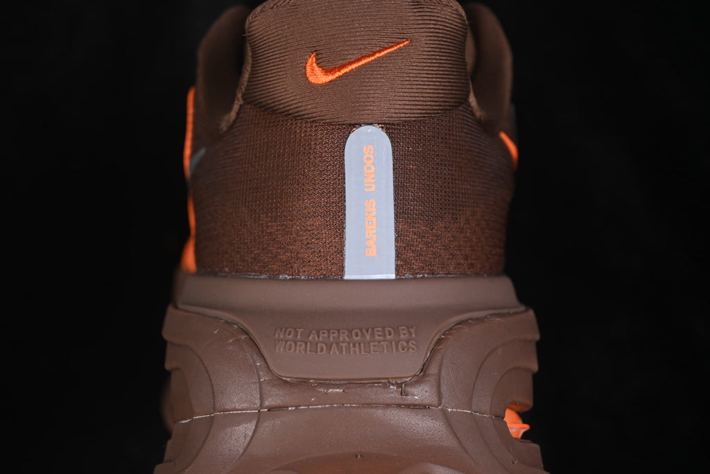 Rear view of the brown sneaker showing the orange Nike logo, vertical heel detail, and the molded text 'NOT APPROVED BY WORLD ATHLETICS'.
