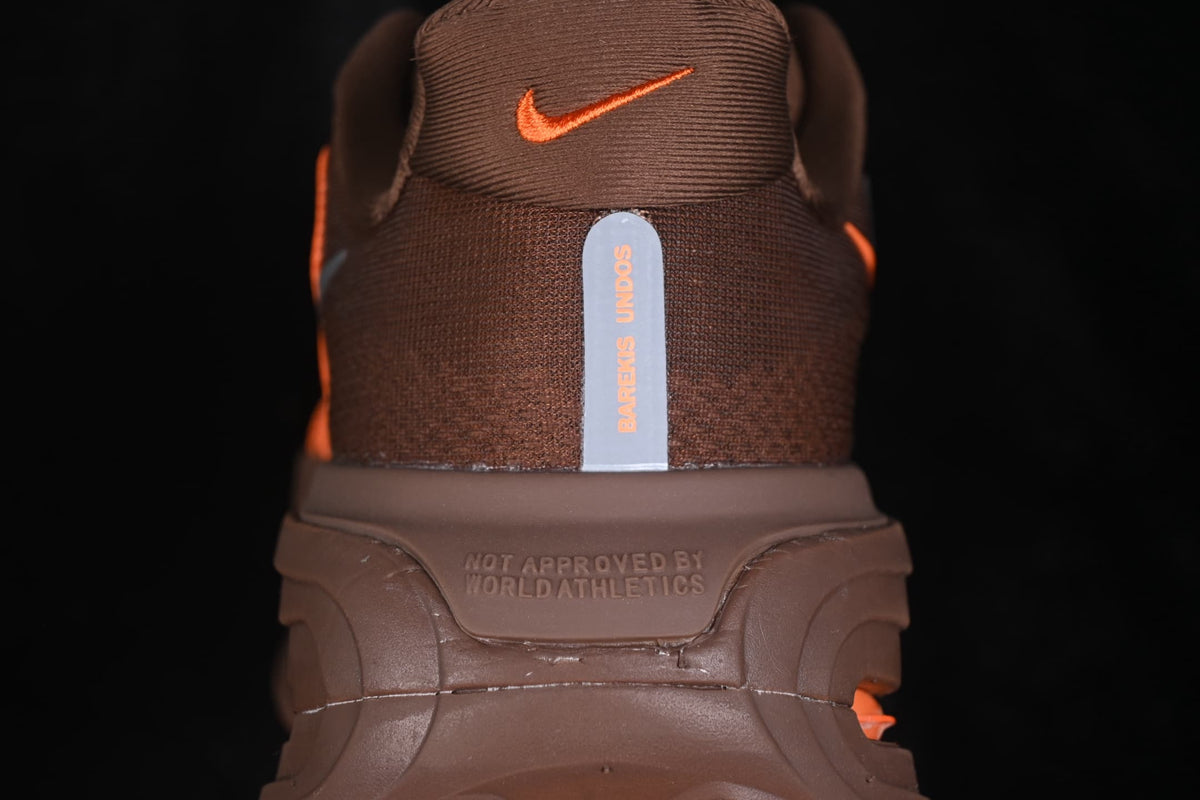 Rear view of the brown sneaker showing the orange Nike logo, vertical heel detail, and the molded text 'NOT APPROVED BY WORLD ATHLETICS'.