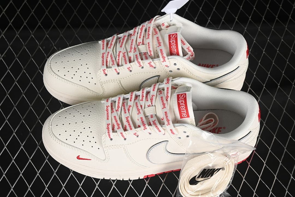 Top view of a pair of cream and red low-top sneakers with Supreme-branded laces and an extra pair of cream laces.