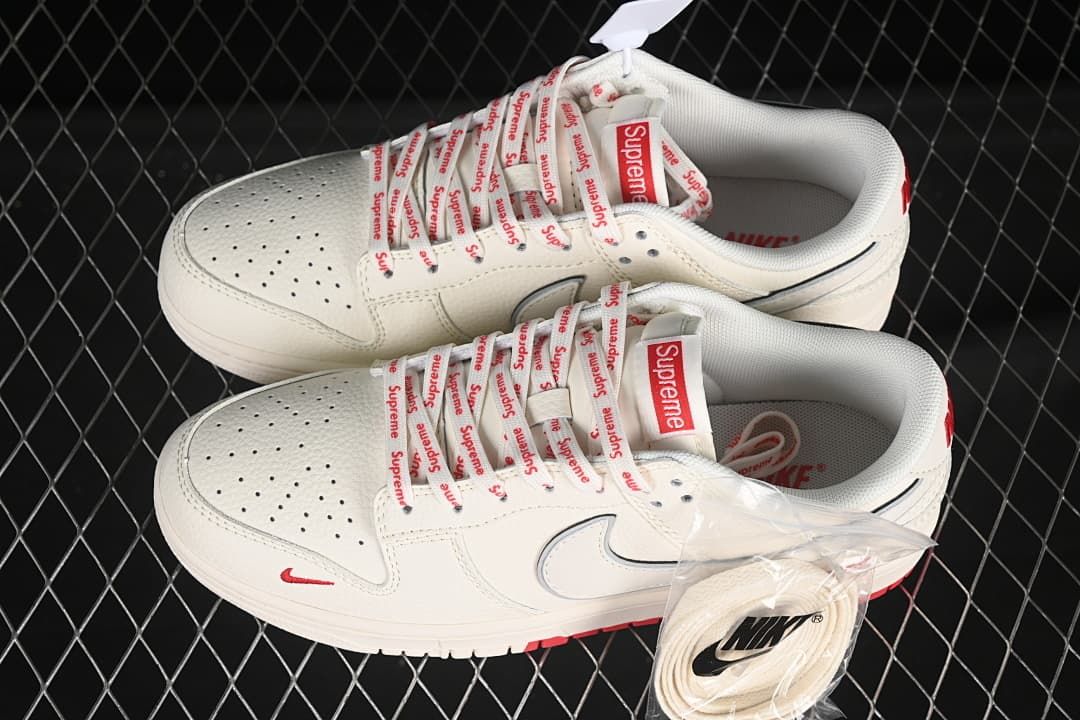Top view of a pair of cream and red low-top sneakers with Supreme-branded laces and an extra pair of cream laces.