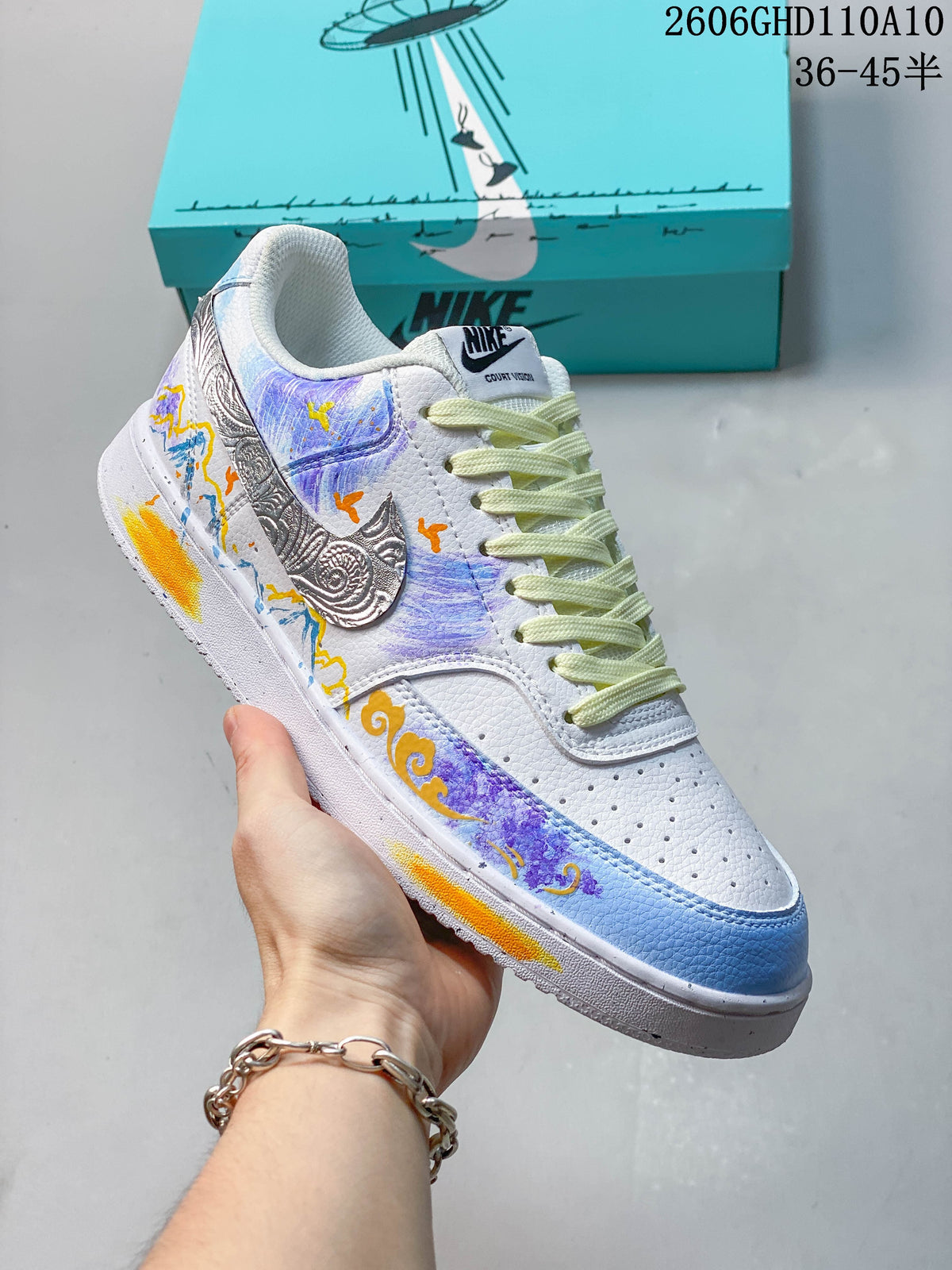 Angled view of a white Nike Court Vision low-top sneaker with blue and purple artwork, orange accents, and a silver-toned patterned side logo.