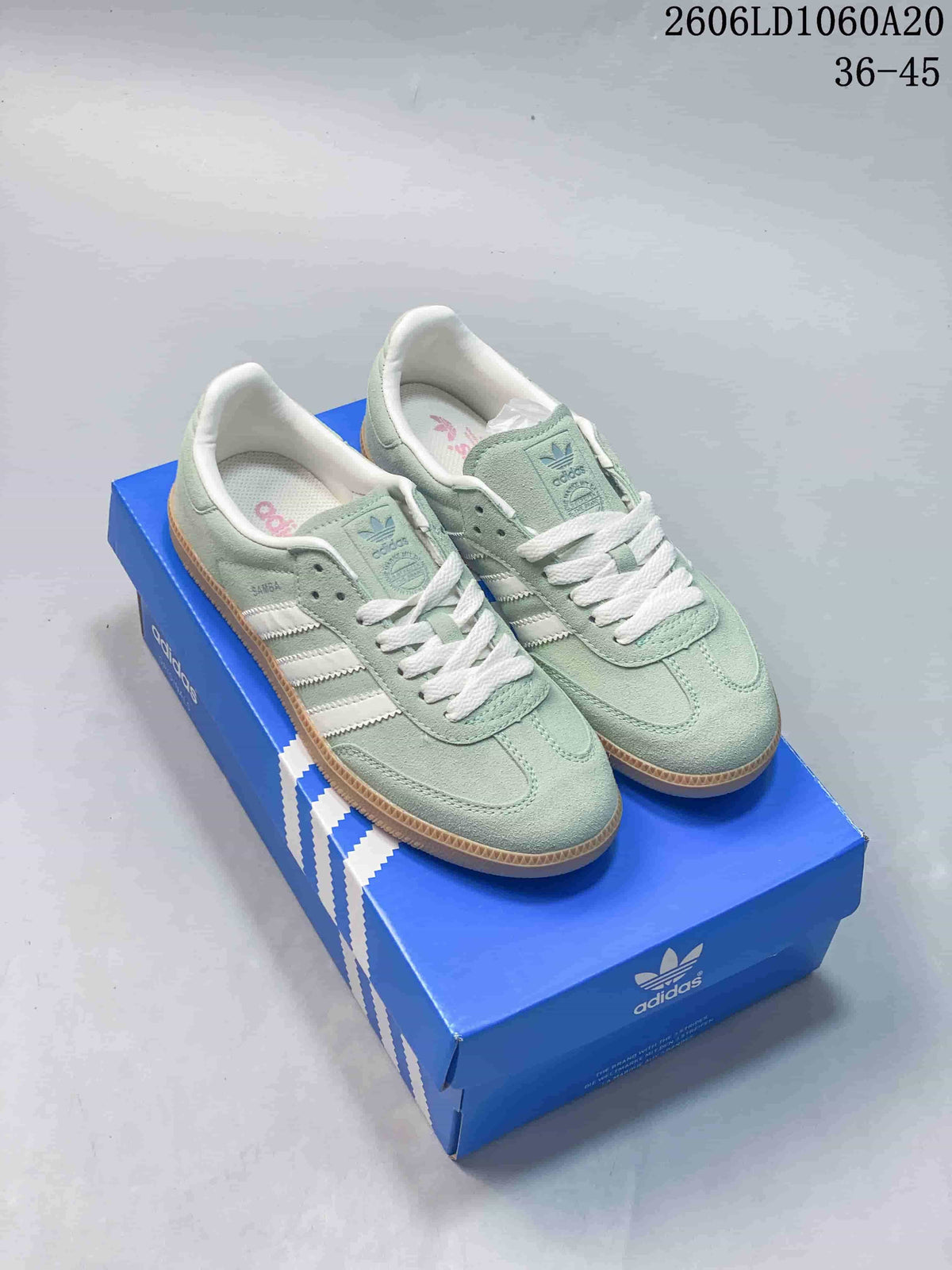 Pair of pale green Adidas Samba OG low-top sneakers with white stripes and gum soles, displayed on a blue shoebox.