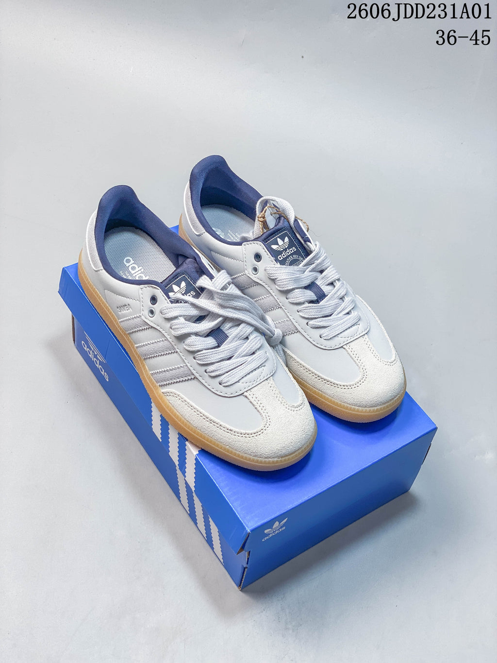 White and gray Adidas Samba-style low-top sneakers with navy accents and gum-colored soles, shown on a blue shoe box.