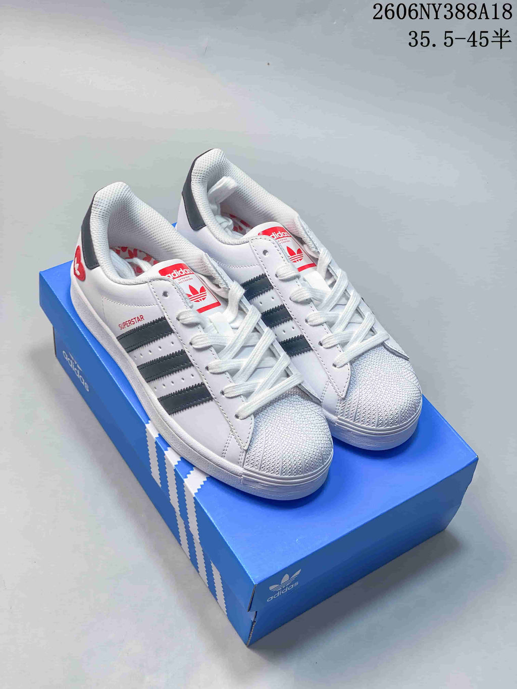 Pair of white adidas Originals Superstar sneakers with charcoal stripes, red details, and textured shell toes