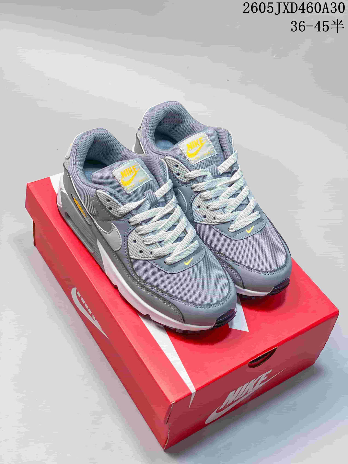 Grey Nike Air Max 90 sneakers with white laces and yellow accents, shown on a red shoe box.