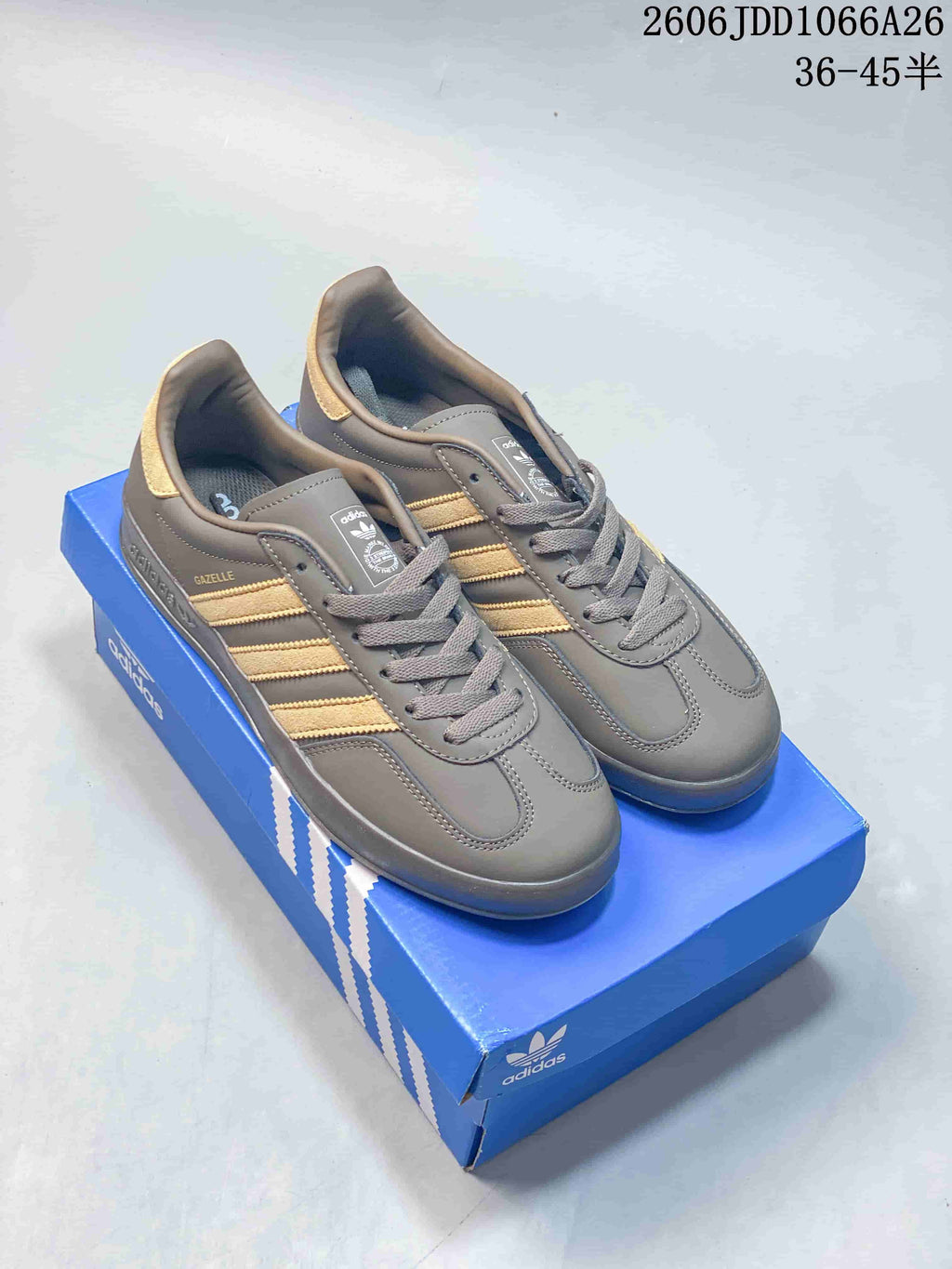 Pair of grey Adidas Gazelle Indoor sneakers with gold-tone stripes, shown on a blue shoe box
