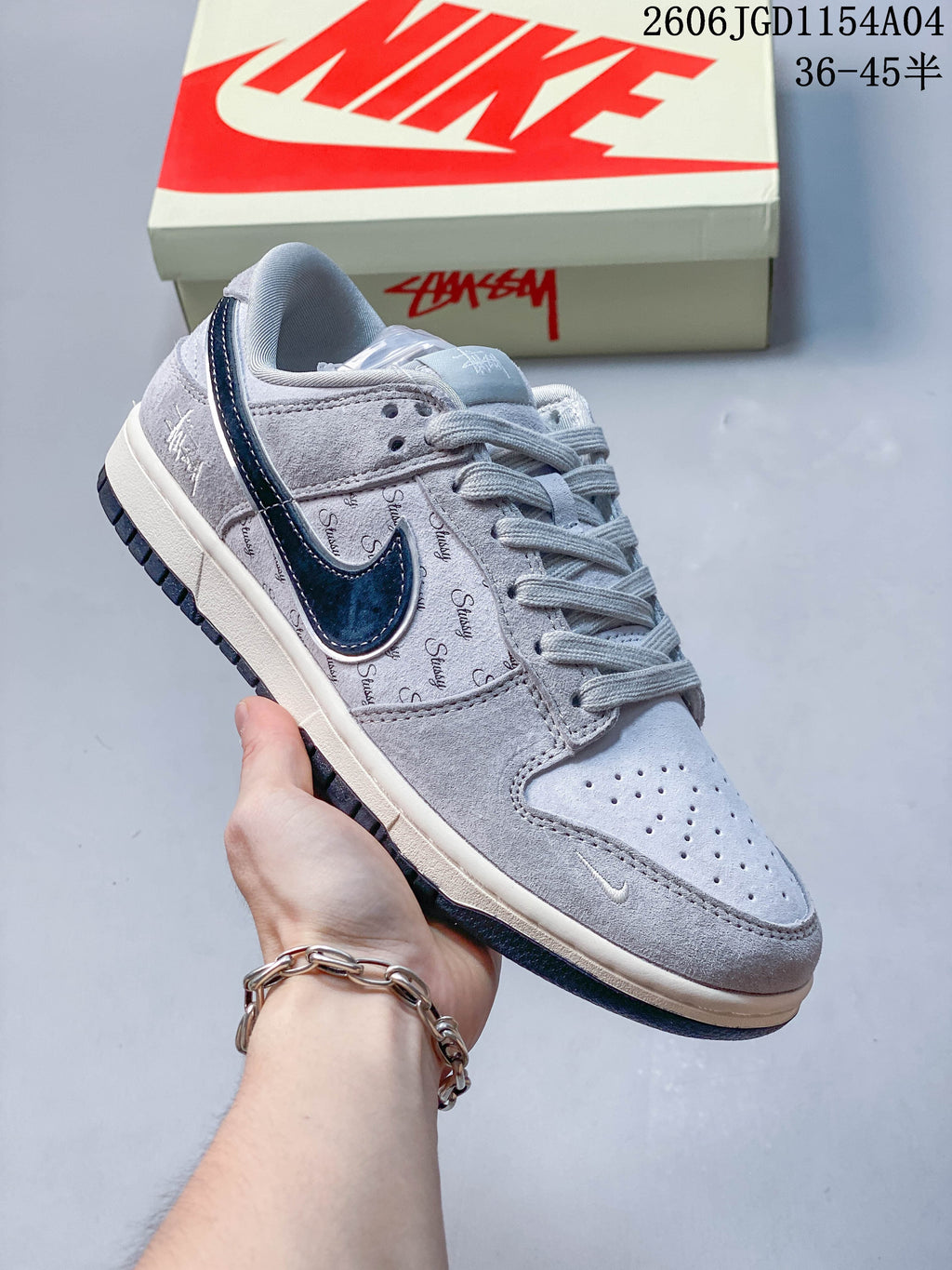 Angled view of a grey and navy low-top sneaker with repeating Stussy script and a two-tone sole