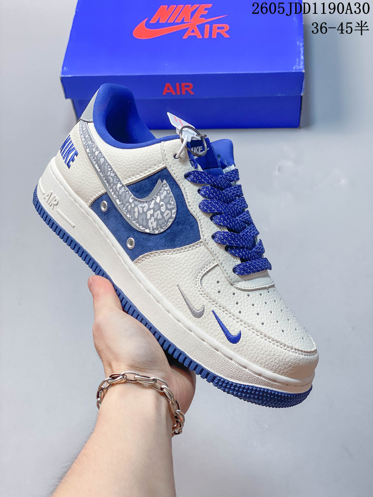 Side view of a white sneaker with blue suede-like panels, blue laces, and a patterned silver swoosh
