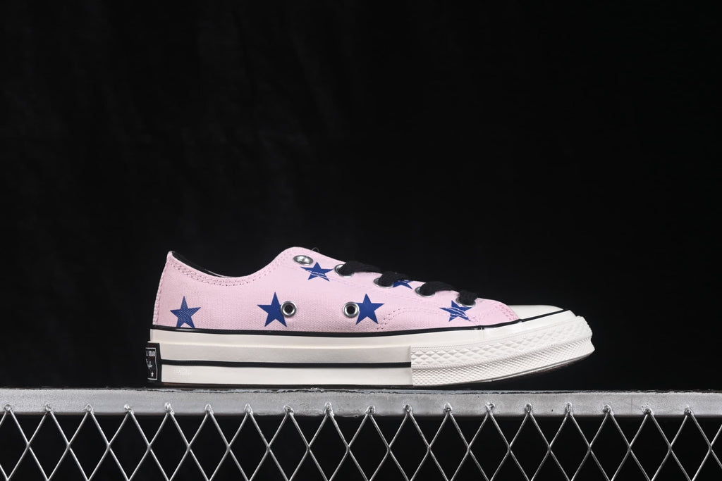 Side view showing the pink upper, blue star pattern, black laces, white toe cap, and layered rubber sole.