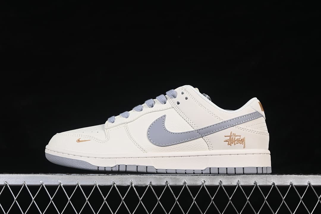 Side view of an off-white Nike SB Dunk Low with a grey Swoosh and gold-tone signature detail.