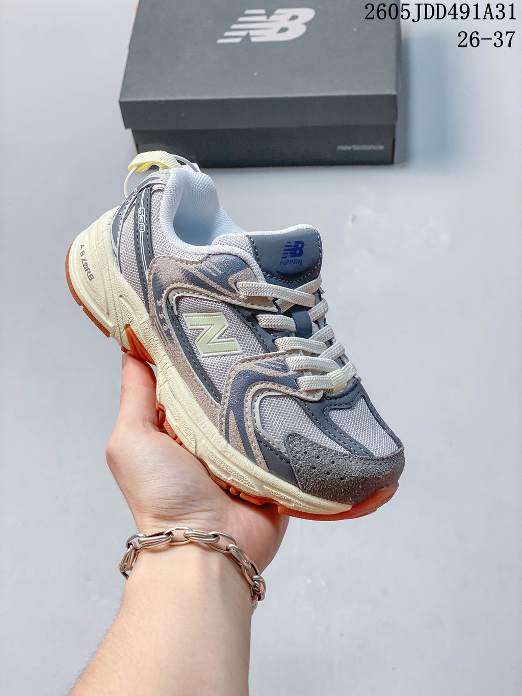 Gray and beige kids' New Balance 530 lace-up sneaker with orange outsole accents, shown in hand.