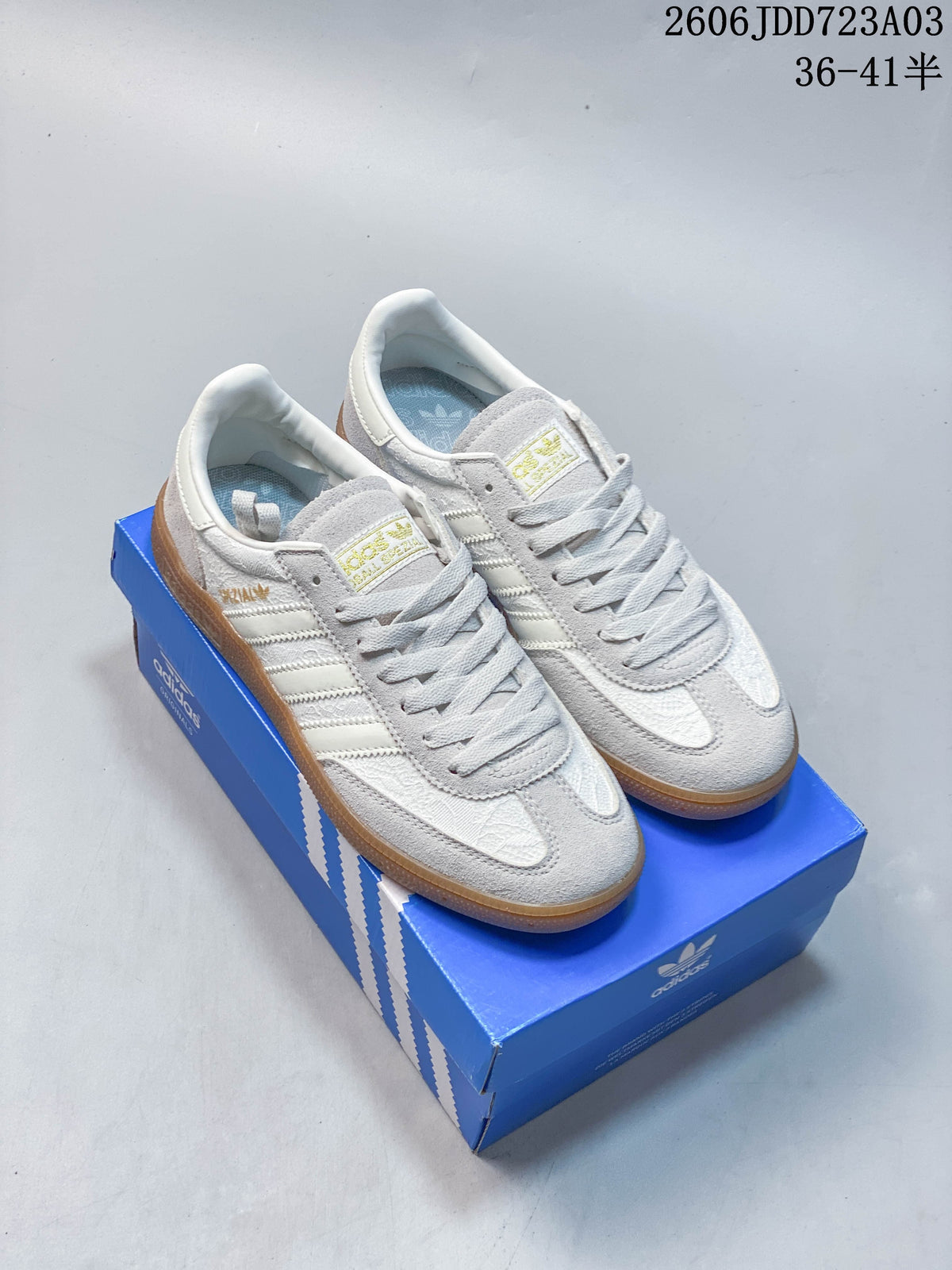 Light gray Adidas Handball Spezial sneakers with pale stripes and a gum-colored outsole, shown on a blue shoe box.