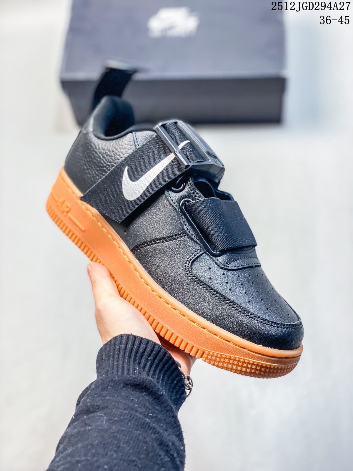 Angled side view of a black sneaker with white Swoosh accent, wide strap, and orange sole