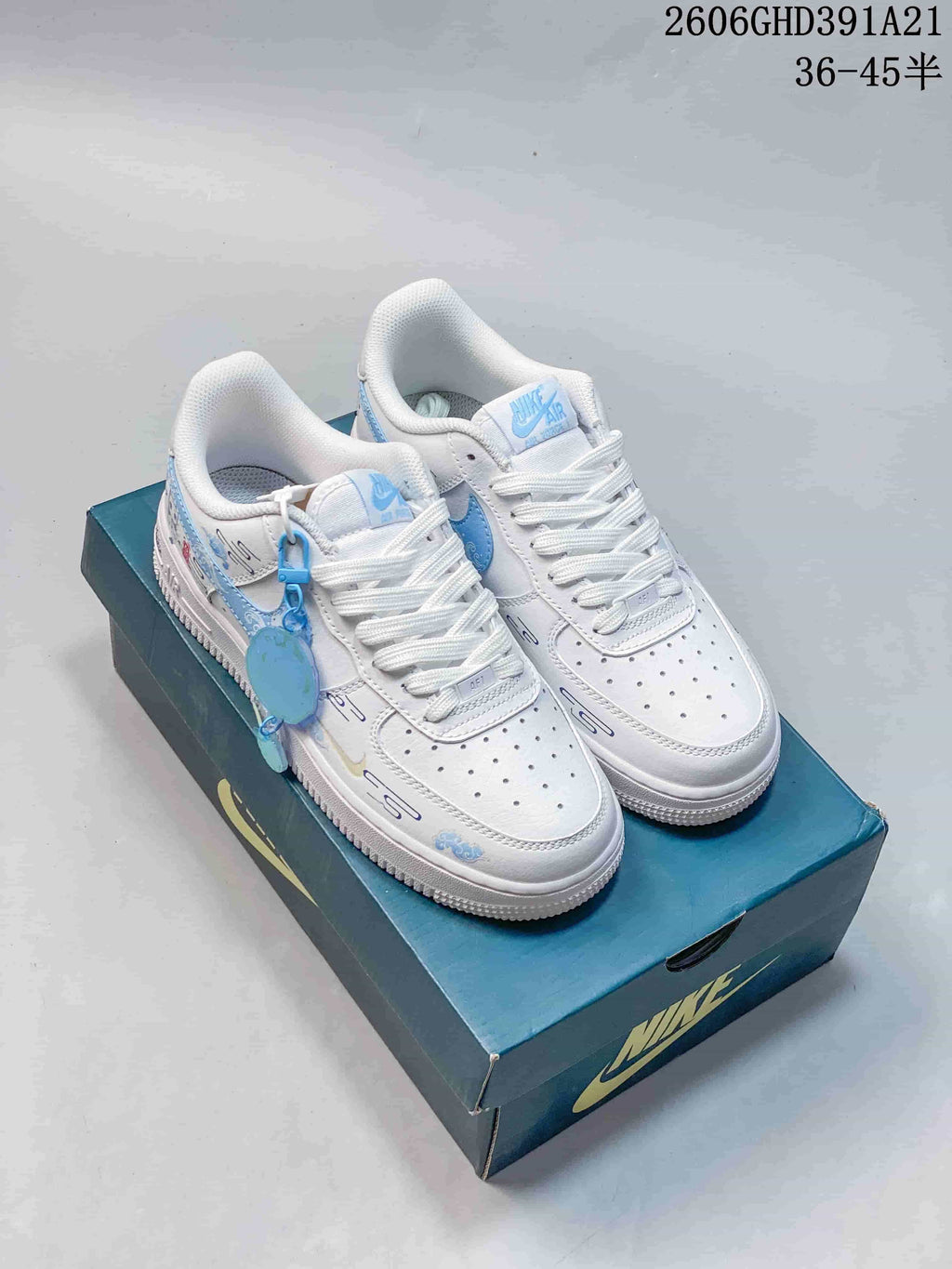 Pair of white and light-blue Nike Air Force 1 Low sneakers with decorative graphics and a blue charm, displayed on a shoebox.