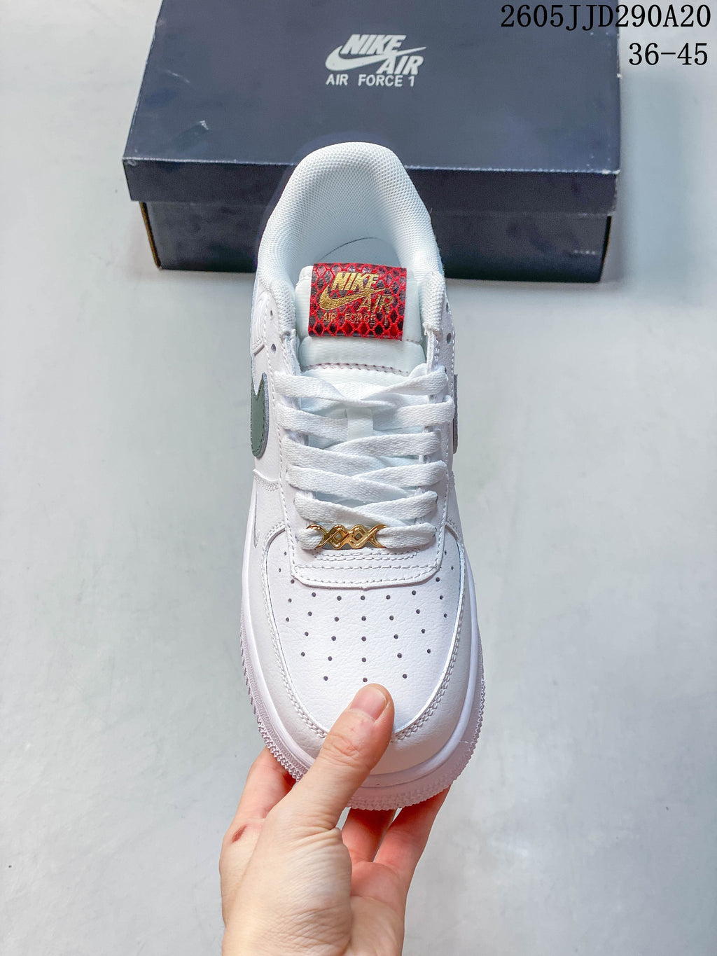 Top view of a white sneaker with perforated toe, red patterned tongue label, and gold-tone lace hardware.