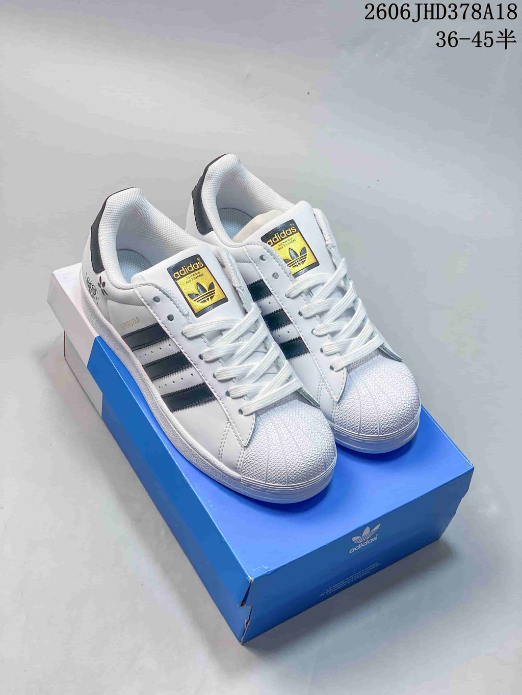 Pair of white Adidas Originals Superstar low-top sneakers with dark side stripes and shell toes, displayed on a blue box.