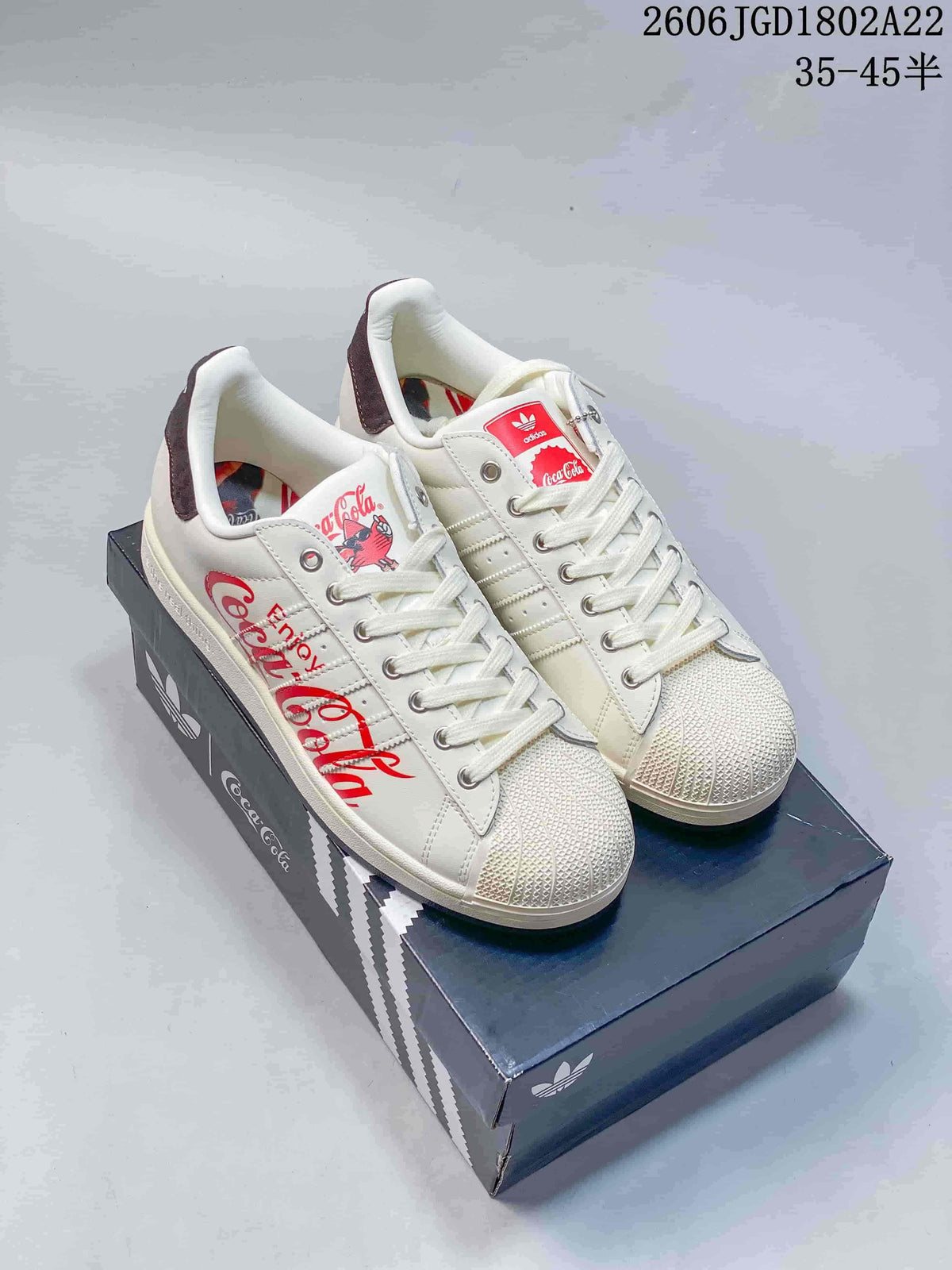 Off-white Adidas Originals Superstar sneakers with red Coca-Cola graphics, shown on a branded box.