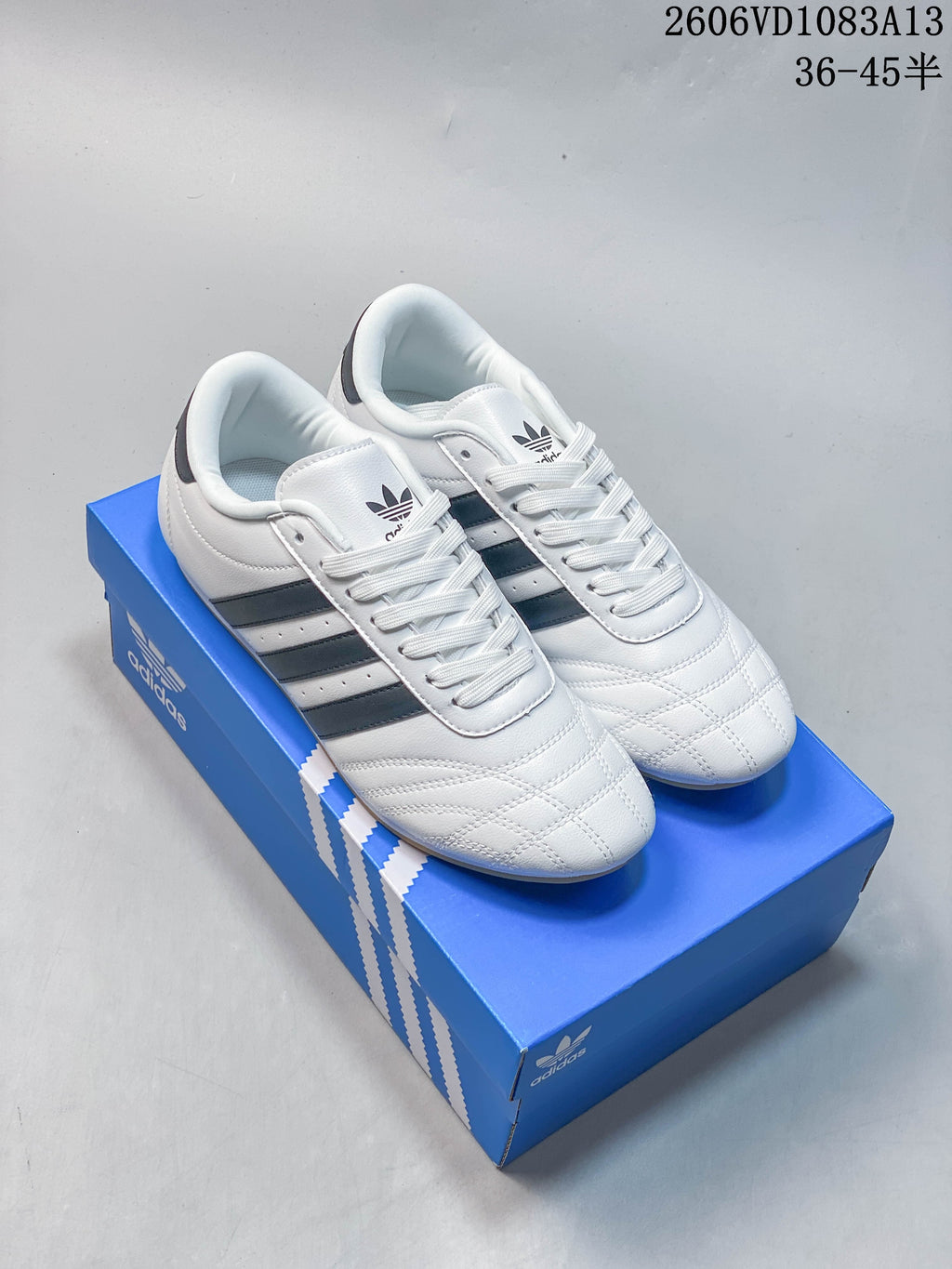 Pair of white Adidas Originals Taekwondo low-top sneakers with dark stripes and laces on a blue shoe box