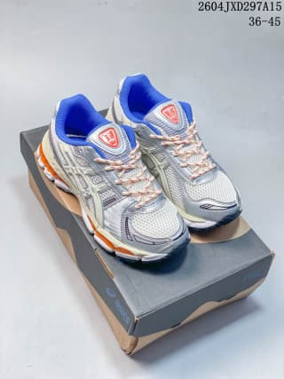 Pair of ASICS sneakers displayed on a shoe box, showing matching light sage, gray, blue, and orange details.