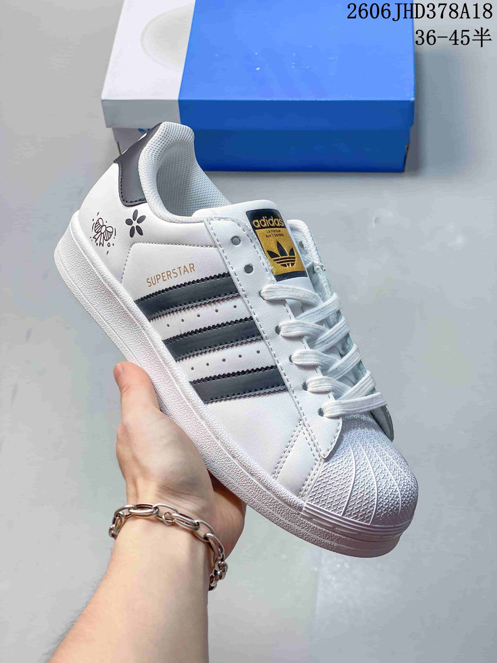 Angled side view showing the white Adidas Originals Superstar sneaker, dark stripes, shell toe, and decorative heel-side graphics.