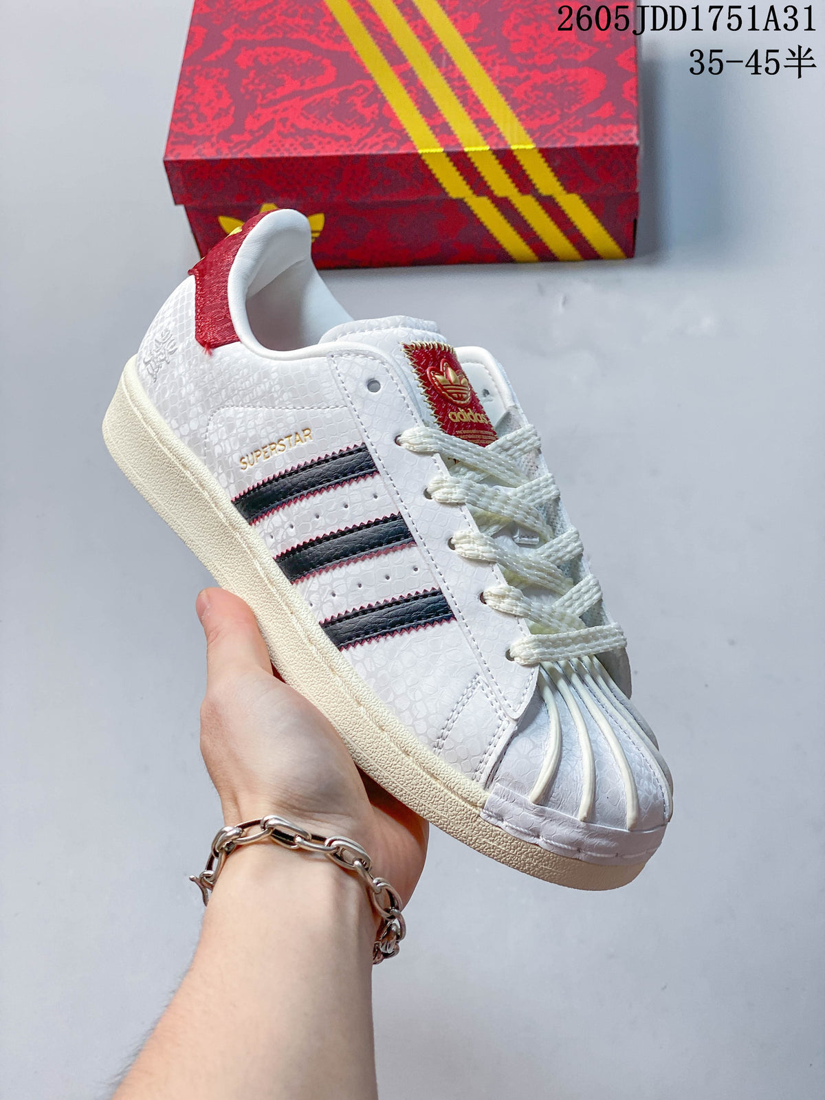 Angled view of the white Adidas Superstar sneaker with dark stripes and red accents