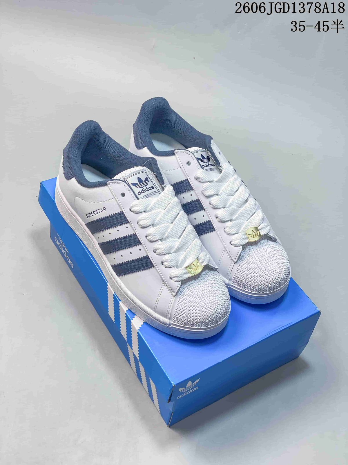 Pair of white Adidas Superstar low-top sneakers with navy stripes and shell-style toes, shown on a blue shoebox.