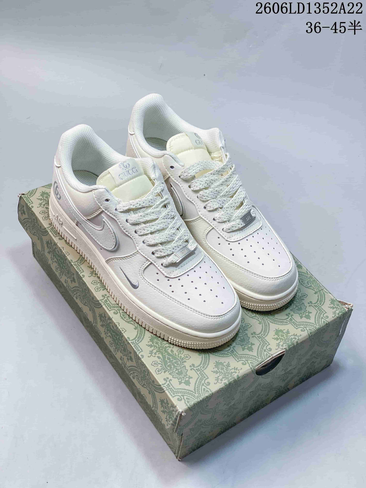 Pair of white Nike Air Force 1 low-top sneakers with pale gray accents on a patterned box; image displays style code and size range.