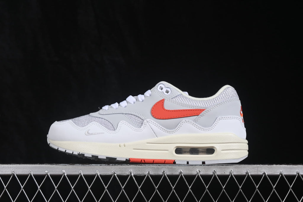 Side profile of a white and grey Nike Air Max 1 sneaker with an orange Swoosh, cream midsole, and visible air unit.